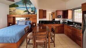 A compact bedroom with a bed, a dining table, and chairs. The room features wooden cabinets, a ceiling fan, and a beach-themed mural on the wall. - Tropical Beach Second Floor Studio with Hill view (Playas Del Coco)