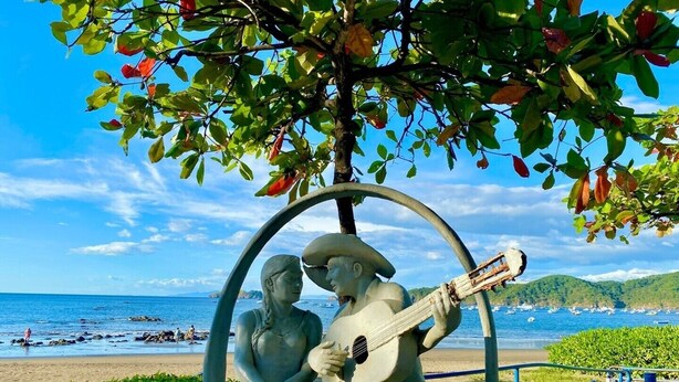 A statue of a man and woman sitting on a bench with a guitar, surrounded by greenery and overlooking a beach. - Tropical Beach Second Floor Studio with Hill view (Playas Del Coco)