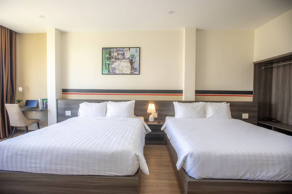 A hotel room with two beds, a desk, and a nightstand with a lamp.