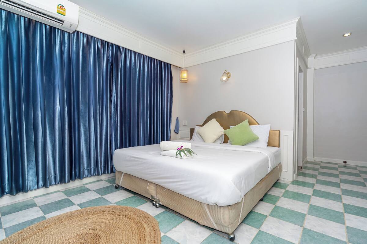 A hotel room with a bed, a large window with blue curtains, a wall-mounted air conditioner, and a checkered floor.