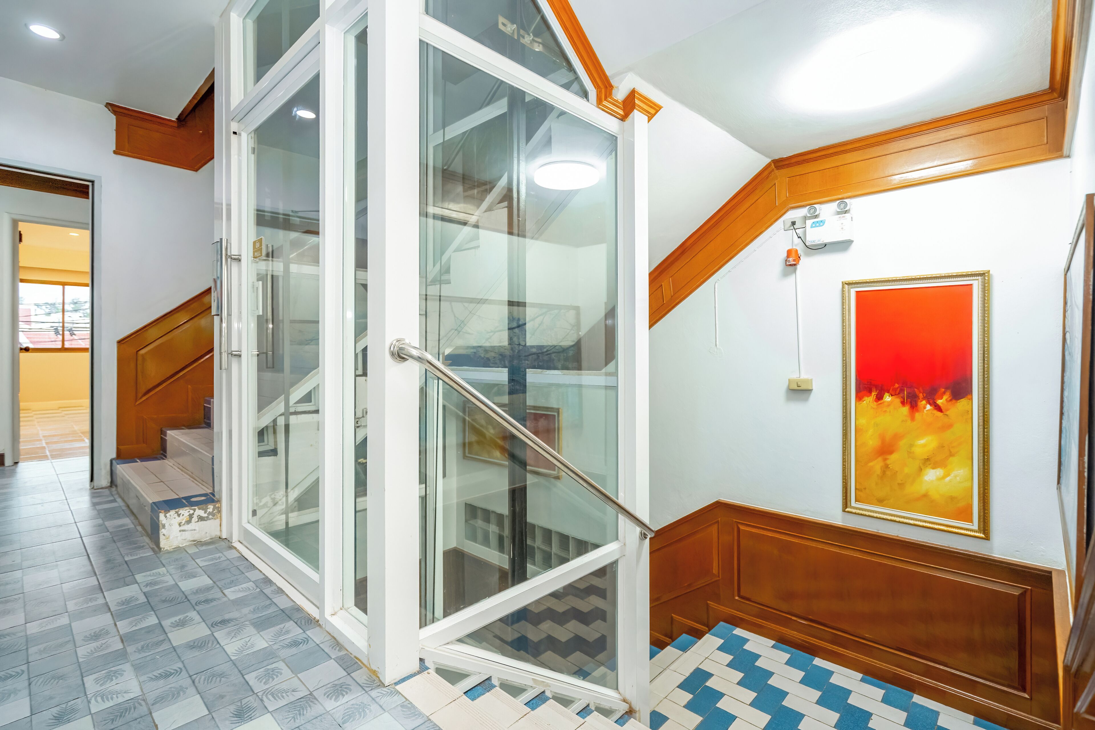 A hotel lobby with a glass door entrance, a staircase with blue and white patterned tiles, and a red framed artwork on the wall.