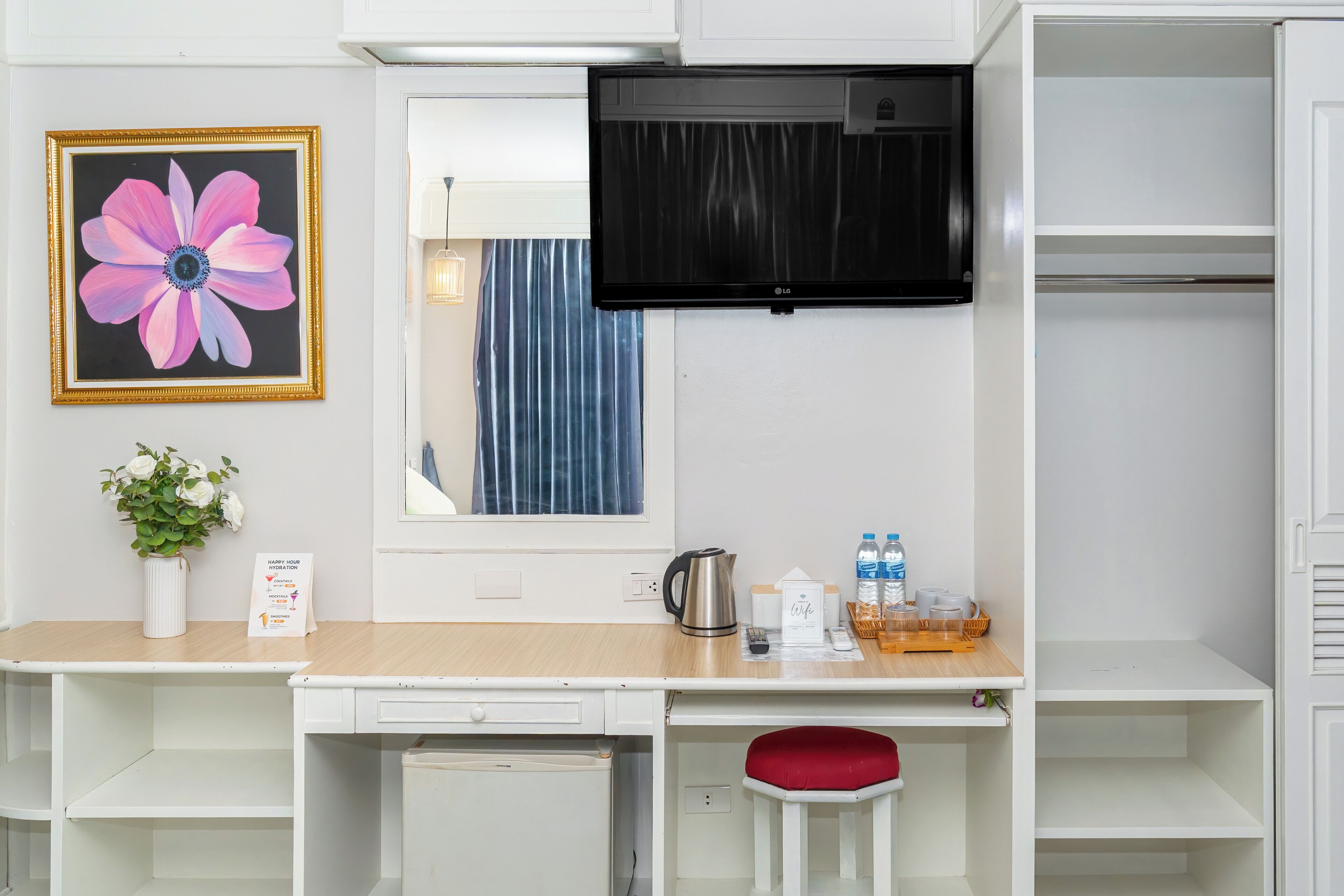 A hotel room with a desk, a mirror, a TV, a red chair, a plant, and a framed flower on the wall.