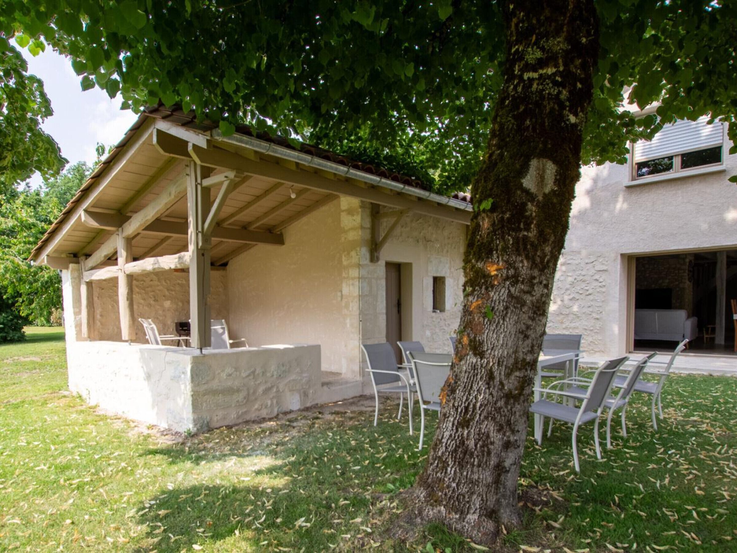 Un espace extérieur aménagé avec des sièges et une table, protégé par un auvent, un bâtiment en pierre doté d’une fenêtre et un arbre au premier plan.