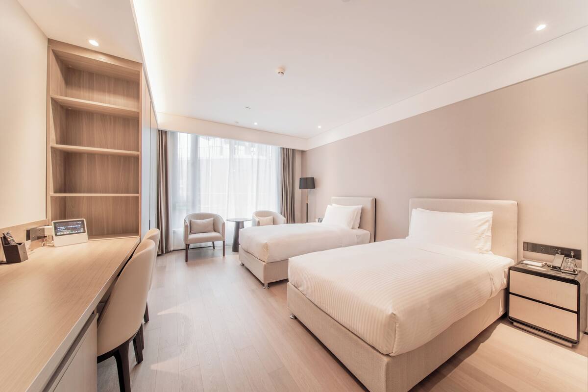 Habitación de hotel con escritorio, dos camas, una silla, una mesita y una mesita de noche con teléfono.