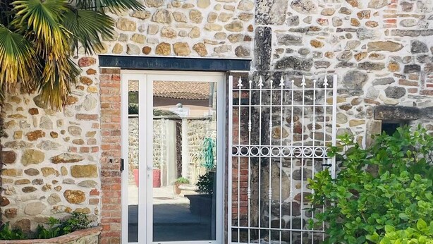 Un bâtiment en pierre avec une entrée fermée par un portail, une terrasse carrelée et des plantes en pot.