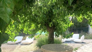 Une table en pierre et deux fauteuils blancs sont placés sous un arbre, dans un coin aménagé en jardin.