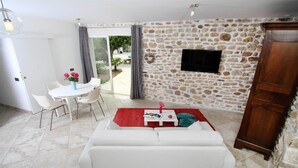 Un salon avec un mur en pierre, un canapé blanc, une table à manger avec des chaises et un téléviseur fixé au mur.