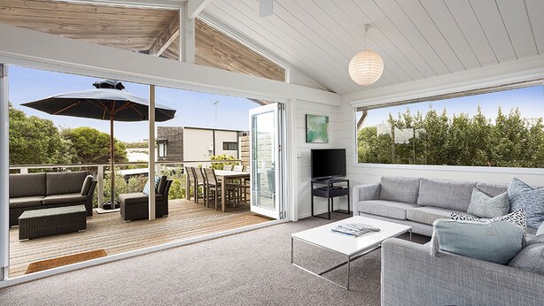Un vaste espace de vie avec une grande porte-fenêtre coulissante donnant sur une terrasse extérieure. L’intérieur comprend un canapé gris, une table basse et un téléviseur.