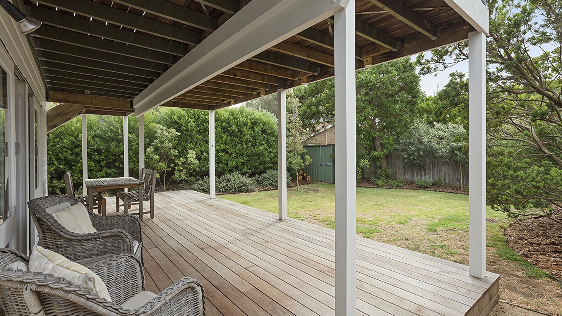 Une terrasse couverte avec un plancher en bois, des meubles en osier et une vue sur un jardin avec des arbres et un abri de jardin.