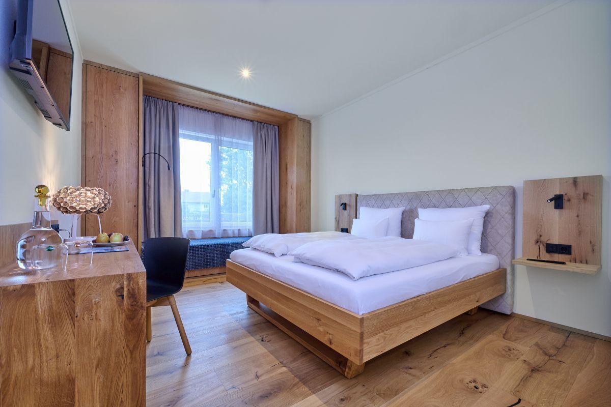 A modern hotel room with a wooden bed, a desk with a lamp, and a large window with curtains.