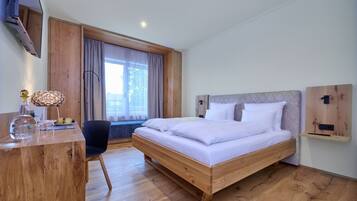 A modern hotel room with a wooden bed, a desk with a lamp, and a large window with curtains.