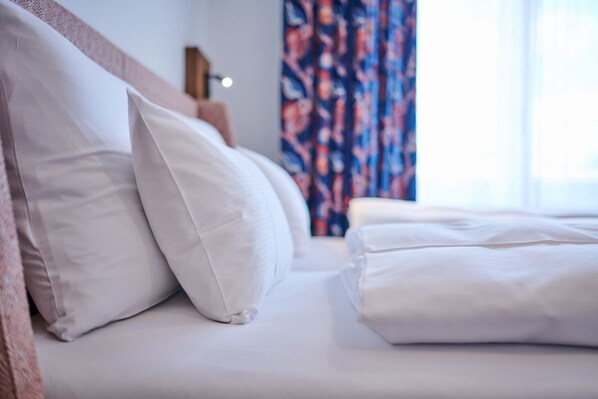 Una cama bien hecha con ropa blanca y almohadas, un cabecero de madera y una cortina floral al fondo.