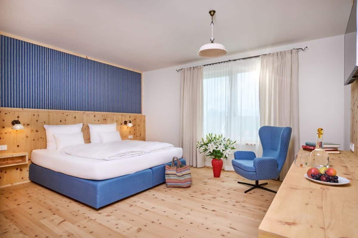 A modern hotel room with a large bed, a wooden headboard, a desk with a chair, a potted plant, and a fruit bowl.