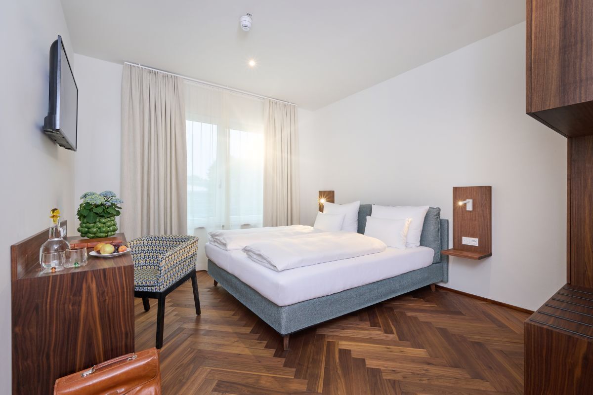 Ein modernes Hotelzimmer mit einem großen Bett, einem Schreibtisch, einem Sessel und einem kleinen Tisch mit Pflanze und Obst.