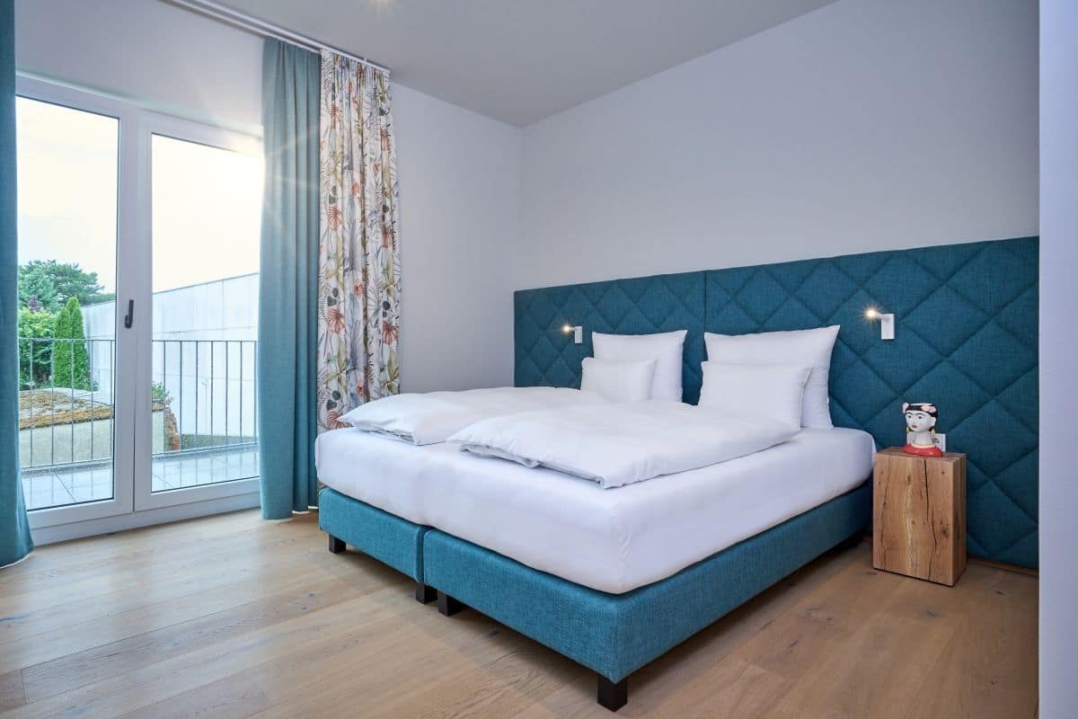A bedroom with a large bed, a wooden nightstand, a blue headboard, and a view of a balcony.