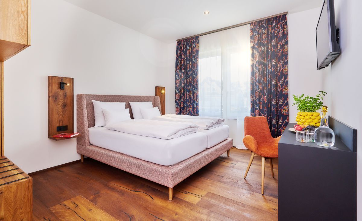 Una habitación de hotel moderna con una cama grande, un escritorio con silla, un televisor montado en la pared y una ventana con cortinas estampadas.