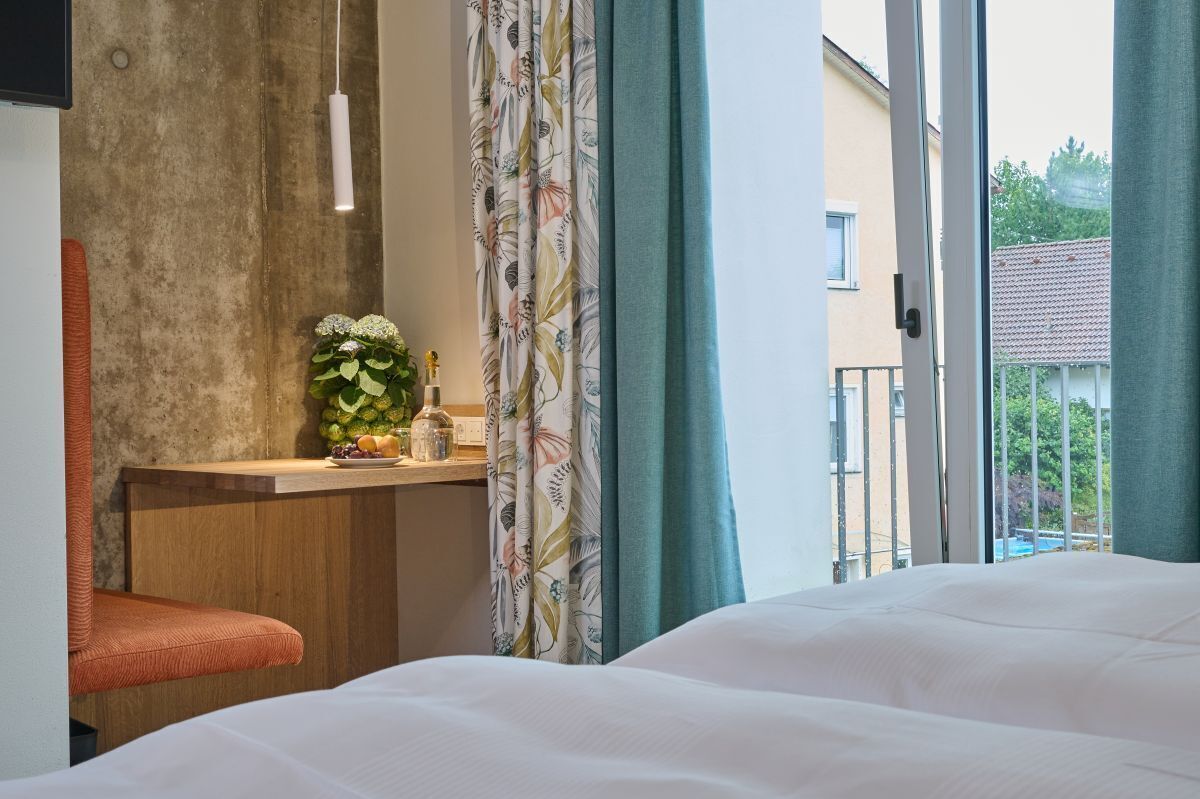 A hotel room with a bed, a small desk with a vase of flowers, a chair, and a view of the outside through a window with curtains.
