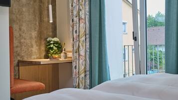 Ein Hotelzimmer mit Bett, einem kleinen Schreibtisch mit Blumenvase, einem Sessel und Blick durch Vorhänge auf die Außenwelt.