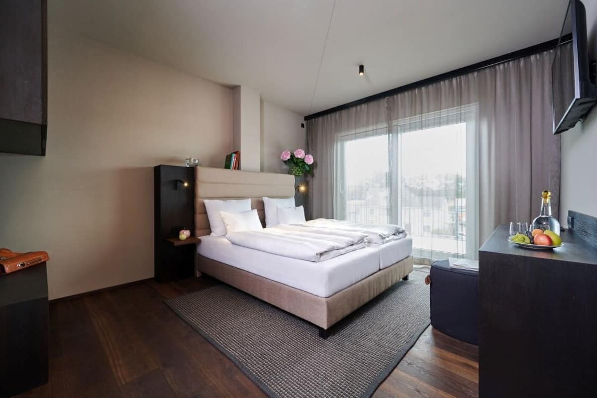 A modern hotel room with a large bed, a desk with a computer, and a view of the city through large windows.