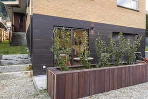 Ein modernes Gebäude mit dunkler Fassade, einer Holzterrasse und einem Pflanzkasten mit Sträuchern.