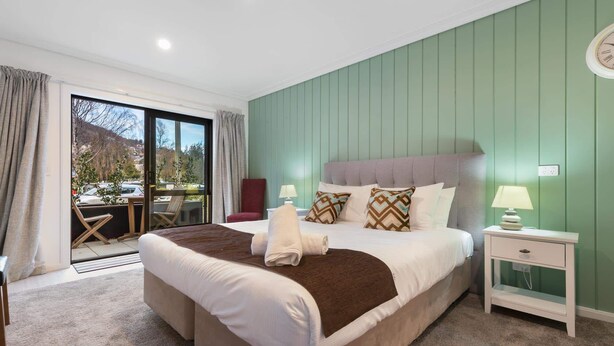 A bedroom with a large bed, a nightstand, a chair, and a view through a sliding door. - Private Studio on Fryer (Queenstown)