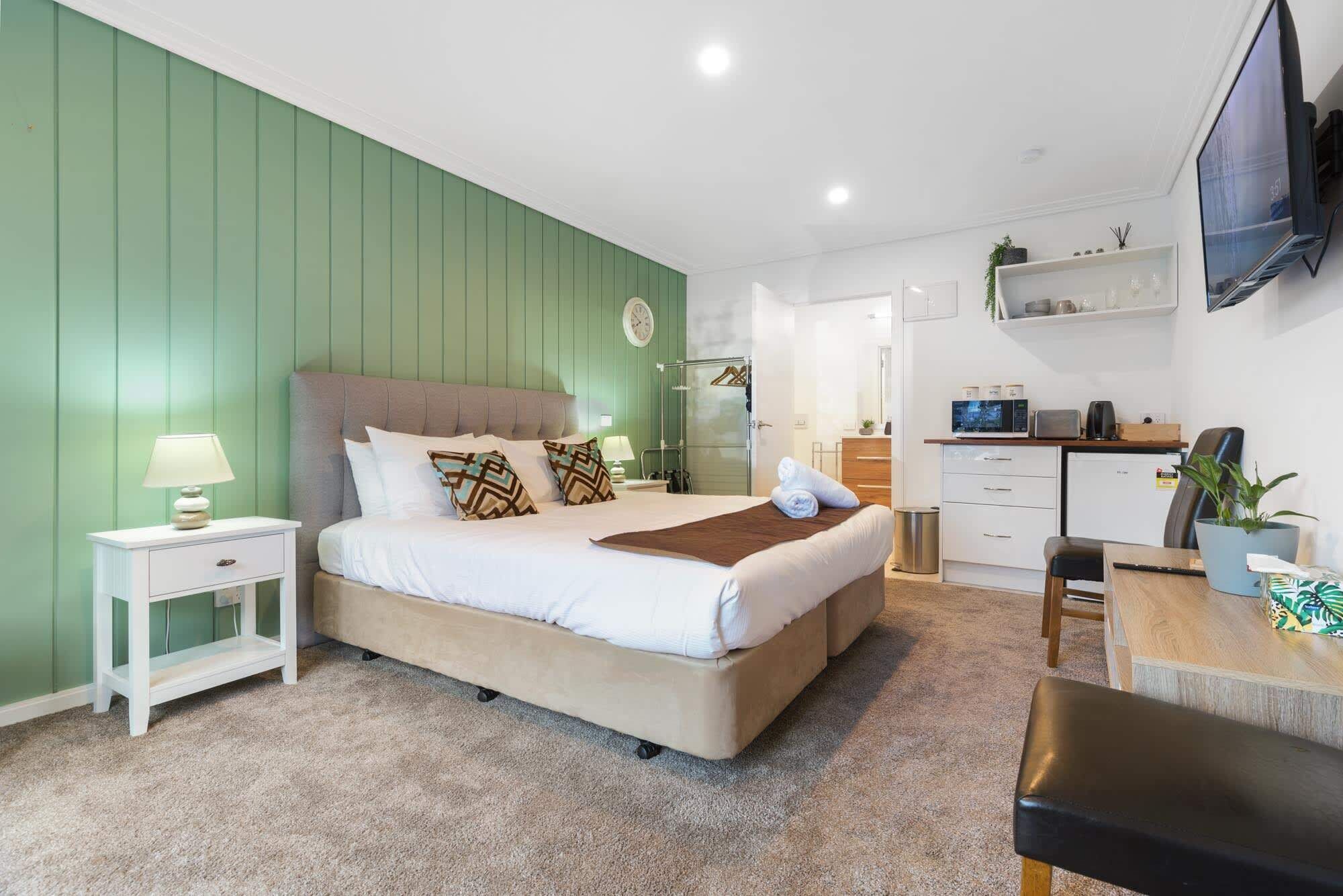 A bedroom with a bed, bedside table, lamp, and a kitchenette area with a TV, microwave, and cabinets.
