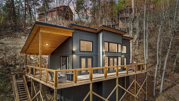 A modern two-story cabin with a wooden deck and stairs, surrounded by trees. - "The Ridgeline" Modern Dog-Friendly Cabin Near PF & Gatlinburg (Sevierville)