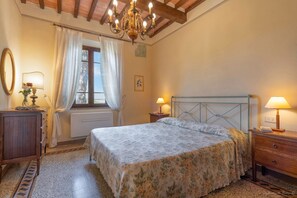 A bedroom with a bed, bedside tables, a radiator, a window with curtains, a chandelier, and a mirror.
