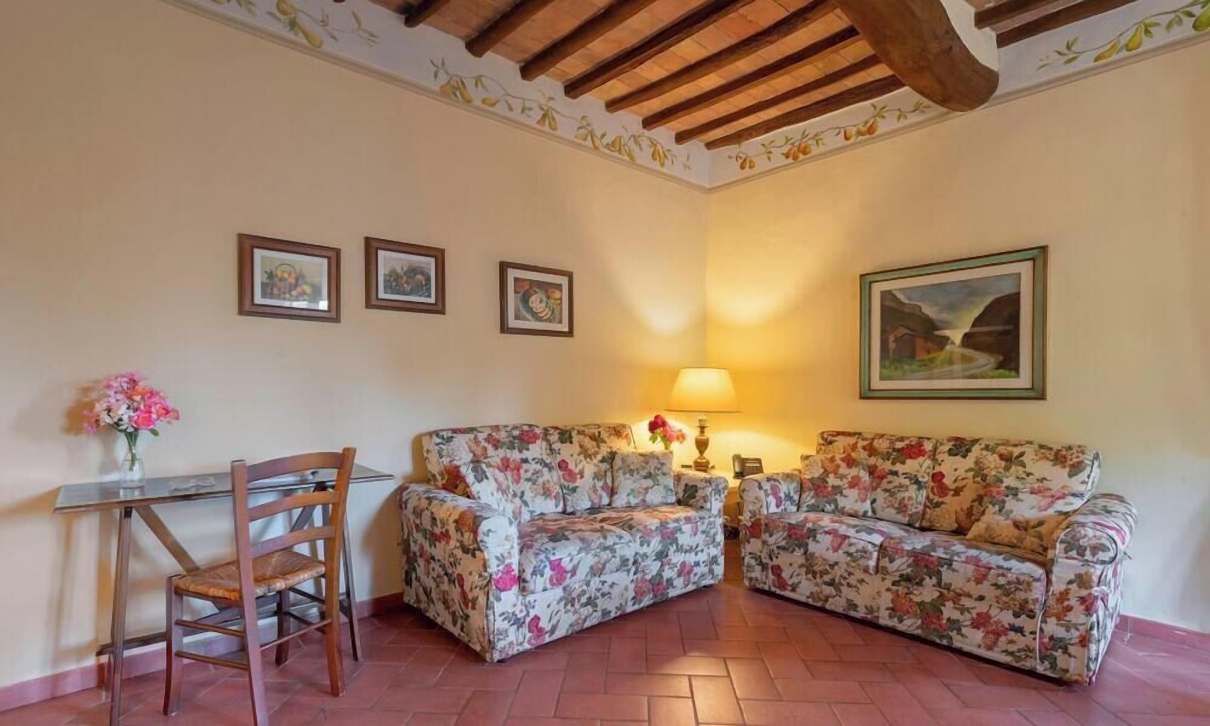 A living room with a wooden ceiling, a floral sofa set, a glass table with a chair, and framed pictures on the wall.