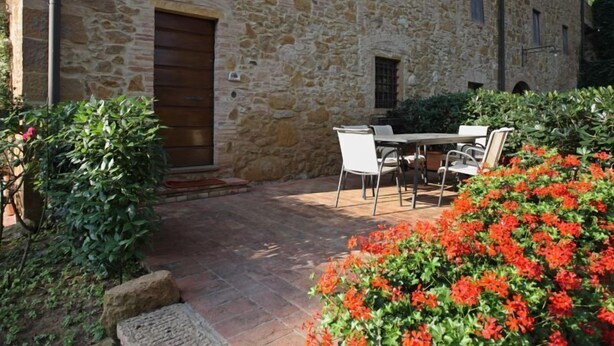 A stone building with a wooden door, a patio with a table and chairs, and red flowers in the foreground. - PINUS - First floor apartment 95 m2 (Montaione)