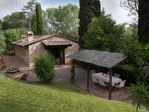 Une charmante maison en pierre avec un toit de tuiles, une terrasse couverte avec une table et des chaises, le tout entouré d’une végétation luxuriante et d’arbres.