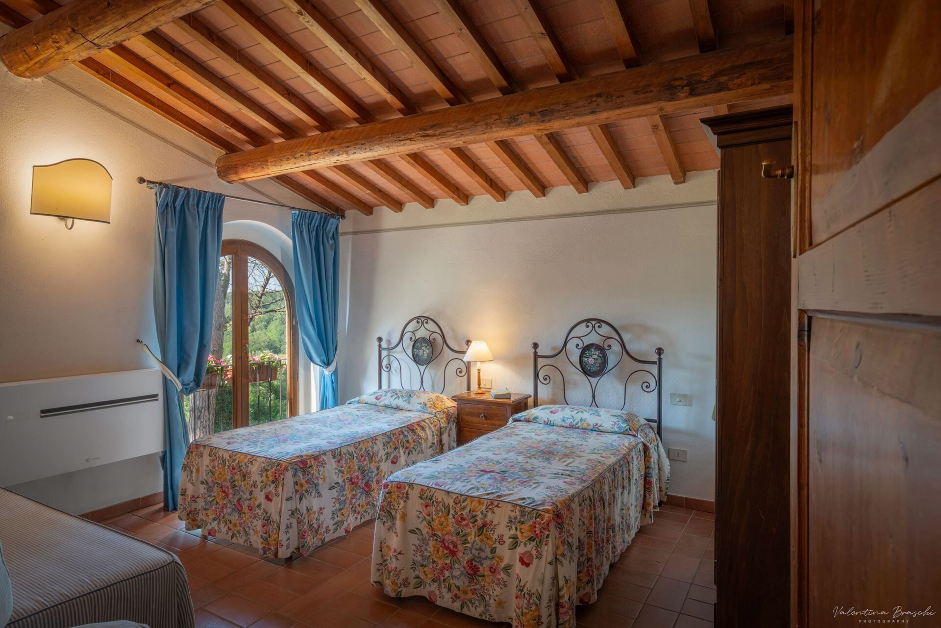 A bedroom with two beds, a wooden ceiling, a window with curtains, and a bedside table with a lamp.