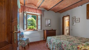 Une chambre avec une porte en bois, une fenêtre donnant sur un paysage verdoyant, un lit recouvert d’une couvre-lit à motifs floraux, une commode en bois et un petit tabouret.