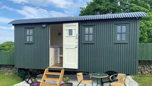Una cabaña pequeña de una sola habitación con la puerta abierta, mostrando una cama y una pequeña cocina americana. Hay una escalera de madera que conduce a la puerta, y una mesa con sillas afuera.