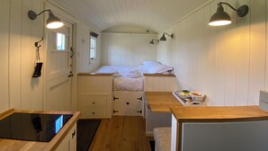 Habitación pequeña y luminosa con cama, una mesita con una bandeja de objetos, estufa integrada y un banco.