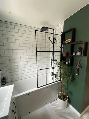 A modern bathroom with a white-tiled shower enclosure, a white bathtub, and a green accent wall with shelves holding decorative items.