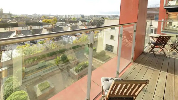 Un balcón con banco de madera y barandilla de cristal, con vista a un paisaje urbano con edificios y vegetación.