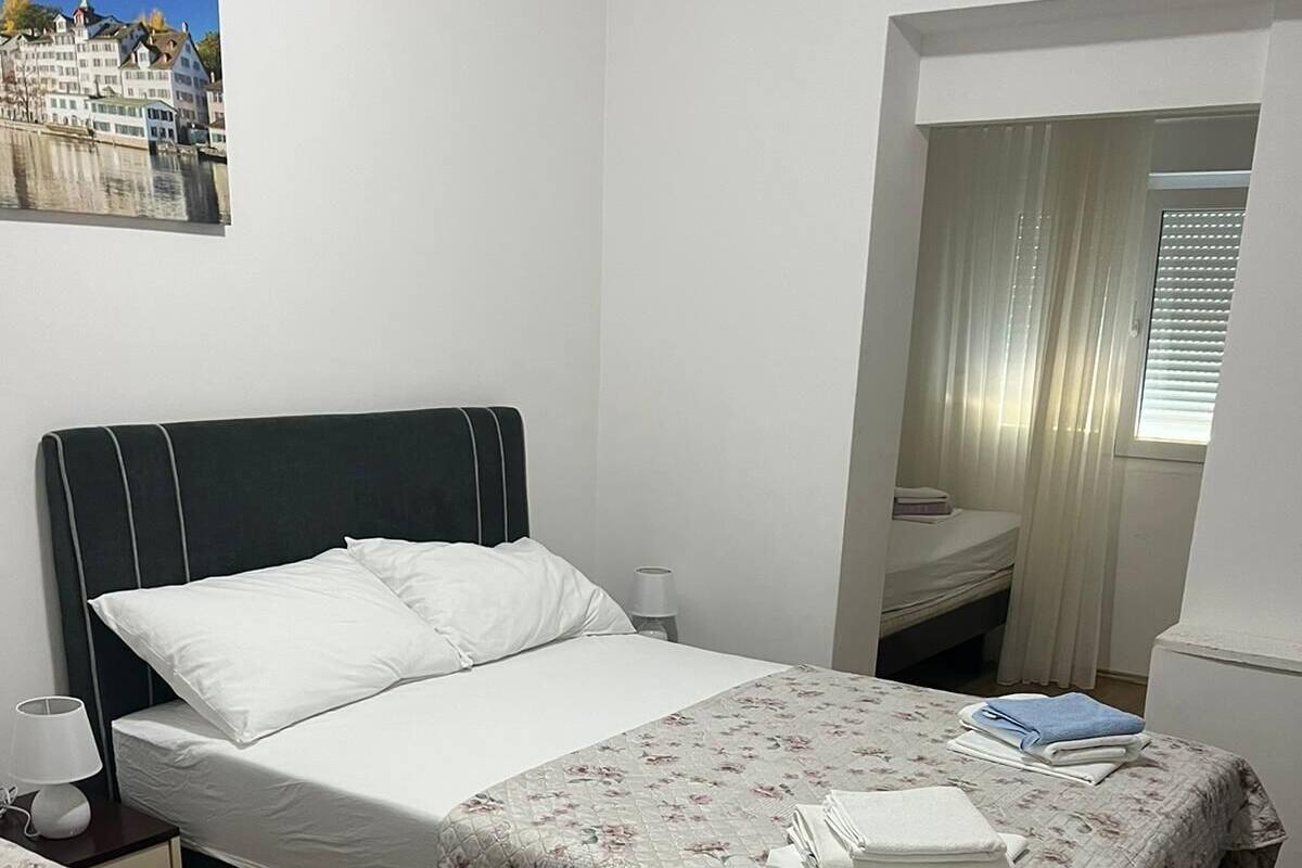 A double room with two beds, each with a floral bedspread and white linens. There is a nightstand with a lamp and a small table with a lamp. A window with curtains is visible, and a painting of a building is on the wall.