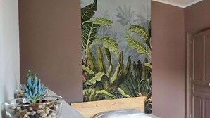 A bedroom with a bed, a nightstand with a plant, and a wall with a tropical mural. - Home for family or business stays (Cambrai)