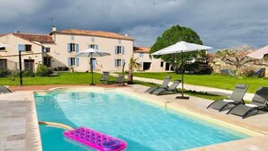 A rectangular swimming pool with a pink inflatable float, surrounded by lounge chairs and umbrellas, with a building in the background.