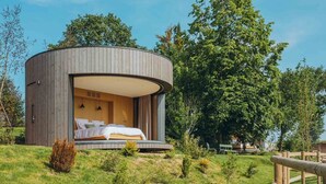 Una cabaña de madera redonda con cama y un pequeño balcón, rodeada de árboles y césped.