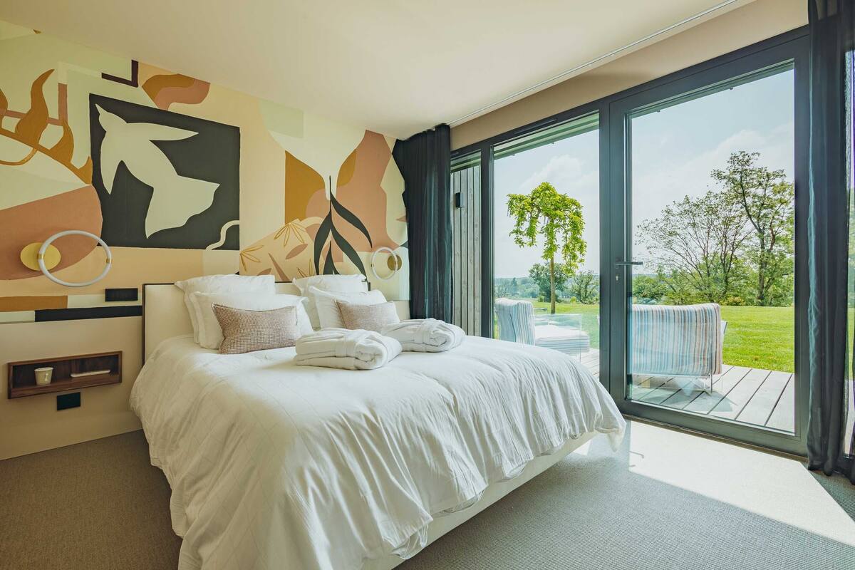 Une chambre moderne avec un grand lit, une vue sur l’extérieur grâce à une porte-fenêtre coulissante et des œuvres d’art abstraites sur les murs.