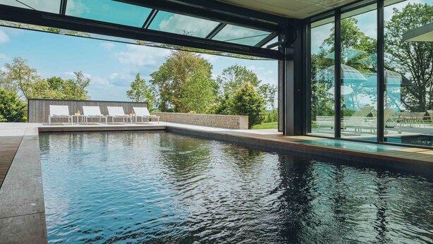Una piscina interior moderna con paredes de vidrio transparente, una terraza de madera y sillones de descanso.
