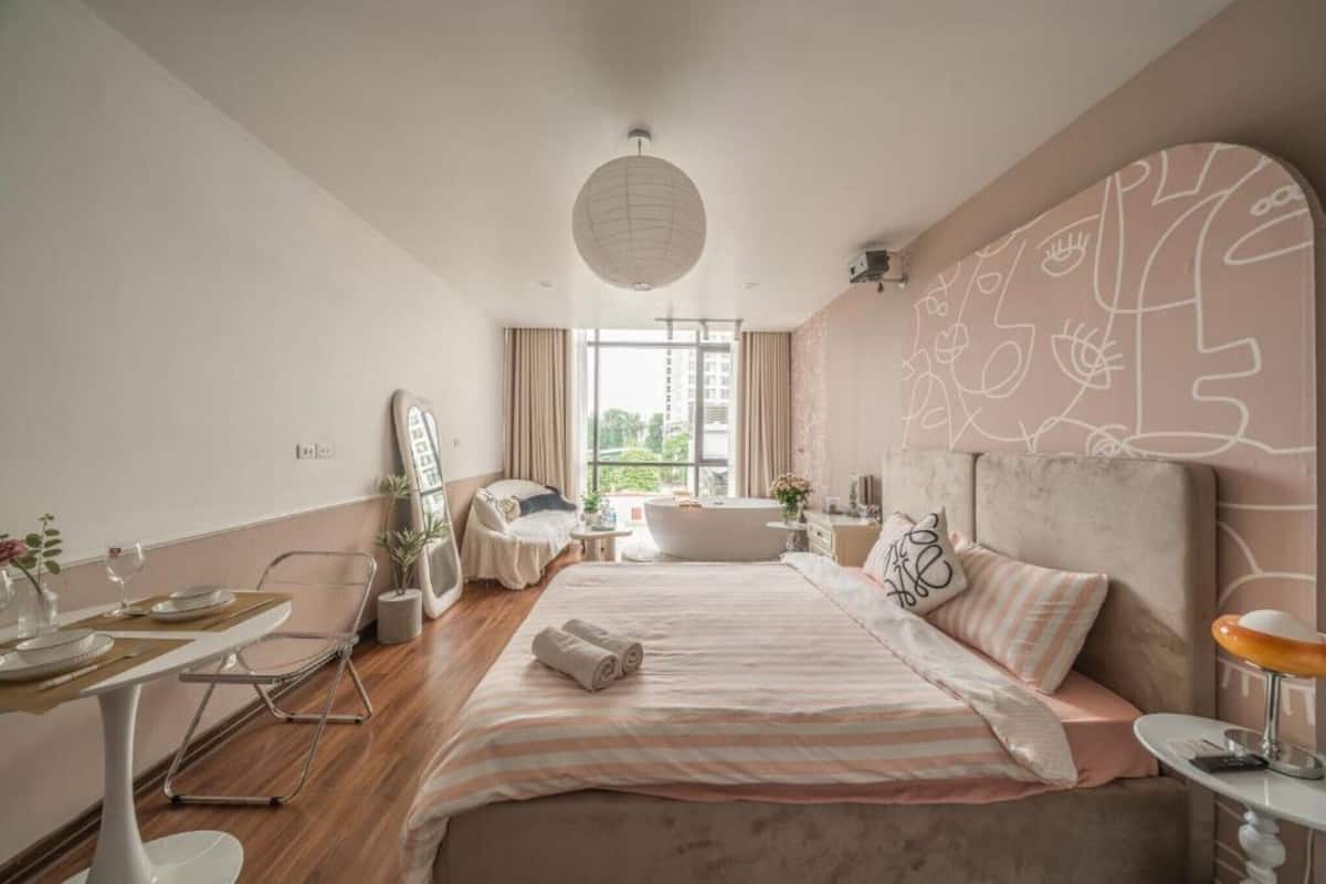 A modern bedroom with a large bed, a sitting area with a table and chairs, a bathtub, and a large window with curtains.