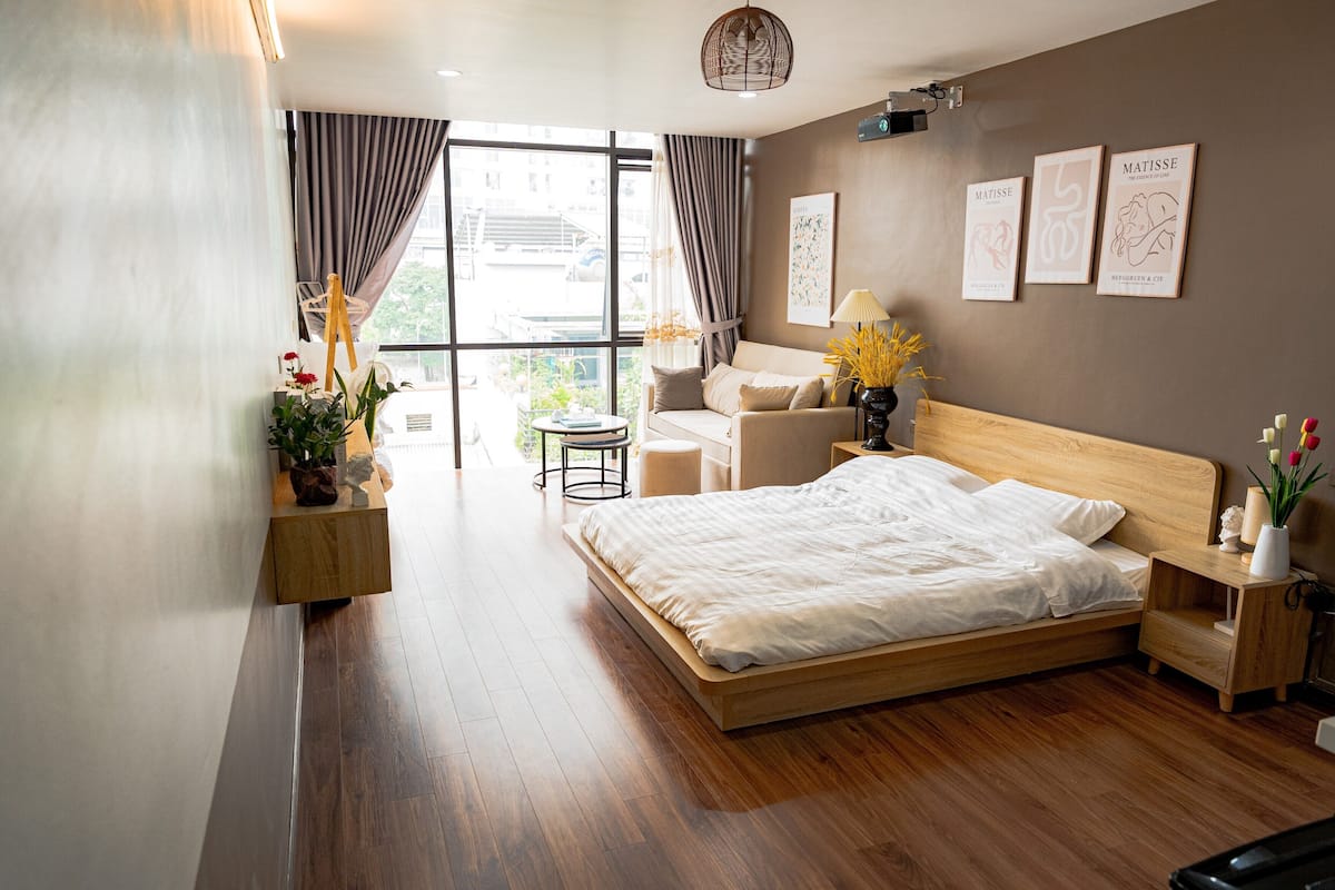 A modern bedroom with a wooden bed, a small round table, a sofa, and a view of the city through large windows.