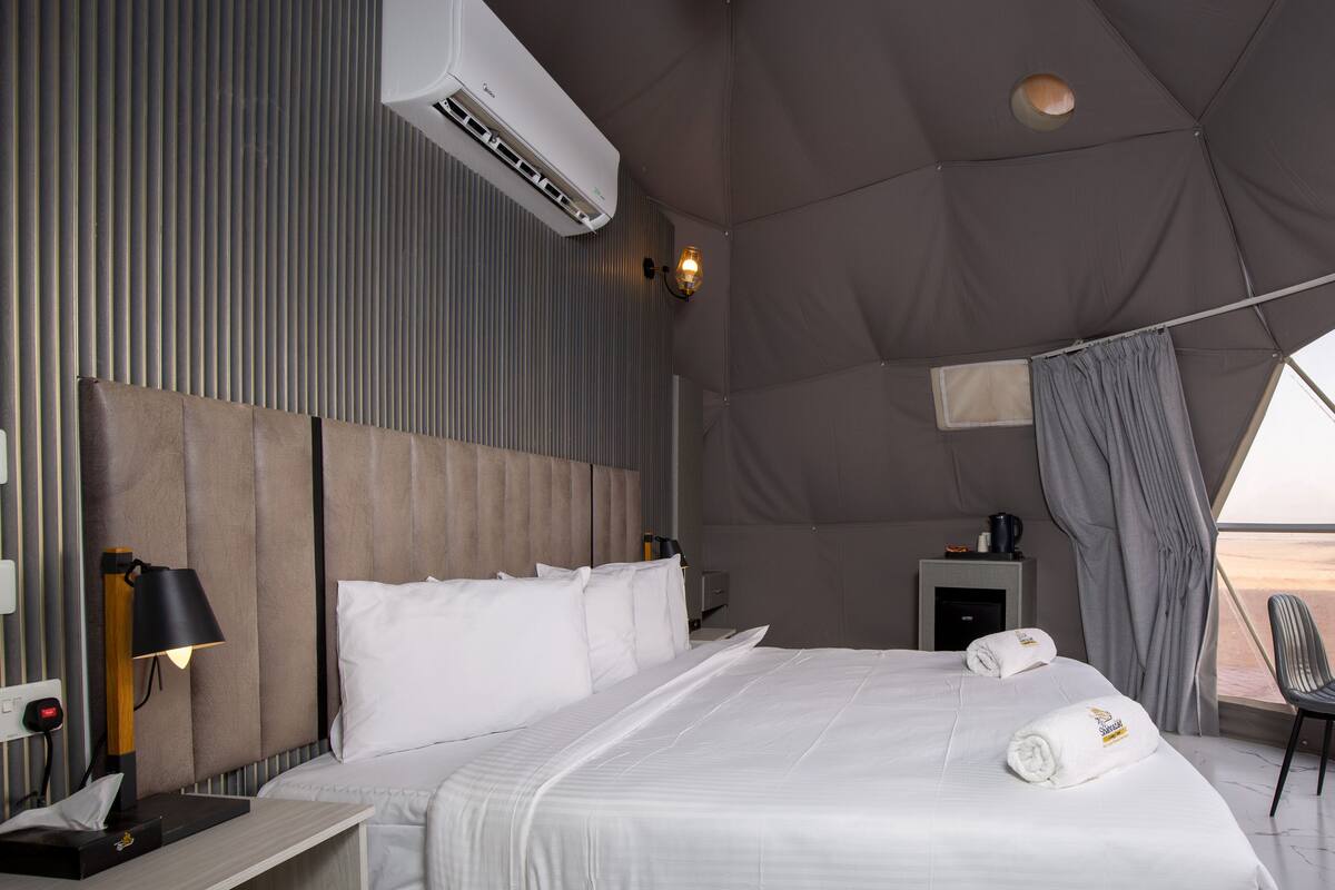 Une chambre d’hôtel moderne avec un grand lit, une table de chevet avec une lampe, une petite table de chevet avec une machine à café, et une vue sur le désert par la fenêtre.