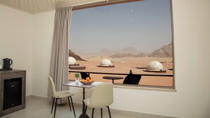 Een kamer met een groot raam dat uitkijkt op een woestijnlandschap met zandduinen en koepelvormige gebouwen. Er staat een klein tafeltje met een schaal fruit en een bloempot, en twee stoelen.