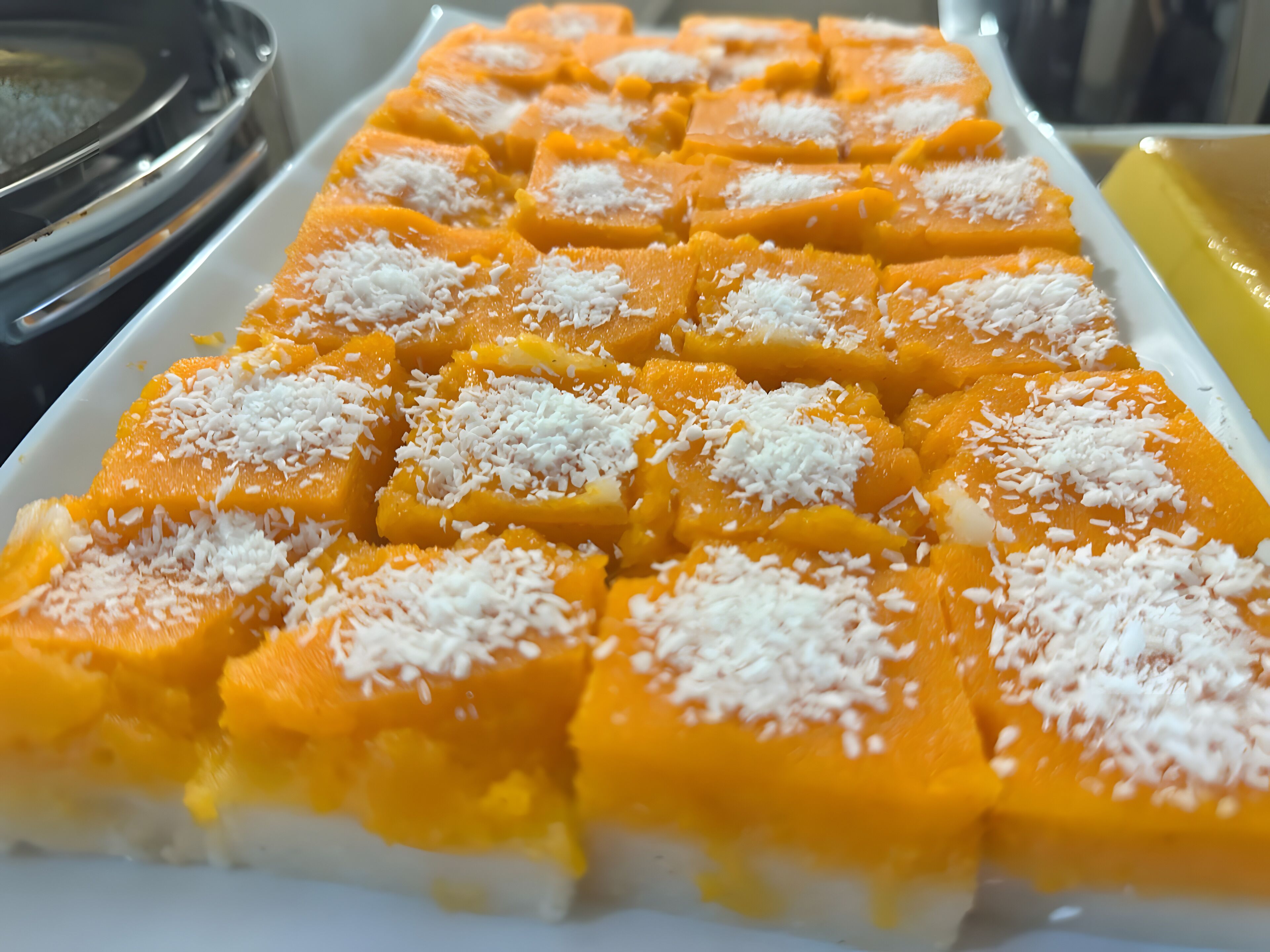 A tray of square-shaped, orange-colored food items topped with white coconut flakes.