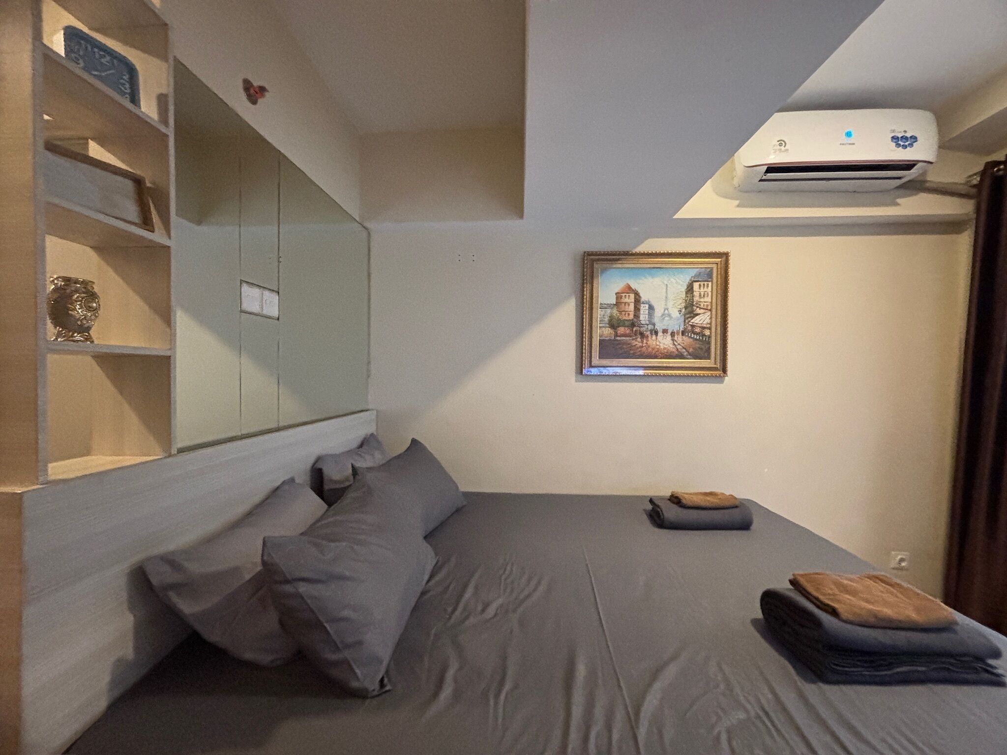 A bedroom with a bed, pillows, a shelf with decorative items, a painting, and an air conditioning unit.