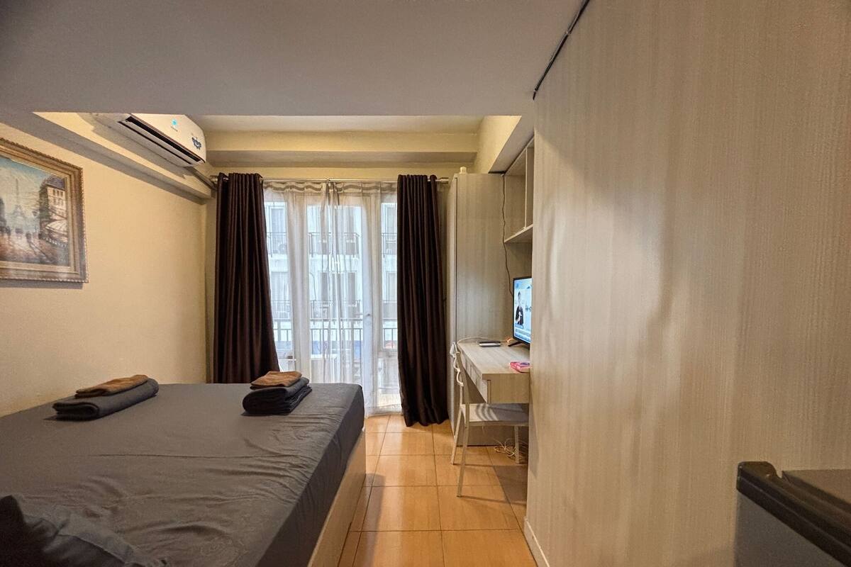 Une chambre d’hôtel avec un lit, un bureau équipé d’un ordinateur, un appareil de climatisation et une vue sur l’extérieur à travers des rideaux transparents.